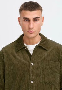 Veste chemise en velours côtelé de couleur vert olive avec un col, une poche avant et des boutons, présentant une texture côtelée verticale et des boutons contrastants.