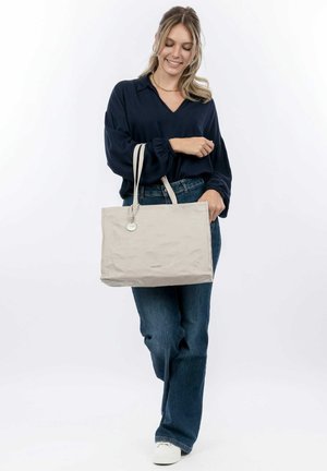 Beige tote bag with a soft texture, two handles, and a decorative tag. The bag features a simple design and is paired with casual denim.