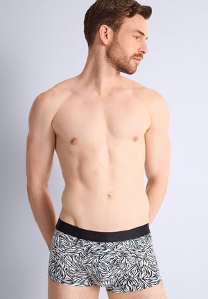 Homme torse nu portant un boxer noir et blanc à motif de feuilles, regardant vers sa droite sur un fond clair uni.