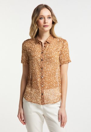 Donna con capelli fino alle spalle che indossa una camicia marrone a maniche corte con bottoni a fiori e pantaloni bianchi, in piedi davanti a uno sfondo uniforme.