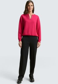 Haut fuchsia à manches longues avec un col en V et de légers plis, associé à un pantalon noir ajusté et des chaussures noires. Tissu lisse, coupe décontractée.