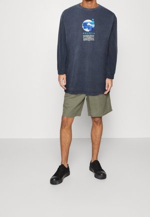 Camiseta de manga larga azul marino con estampado gráfico; combinada con pantalones cortos verde oliva y zapatillas negras; conjunto informal con corte relajado y diseño sencillo.