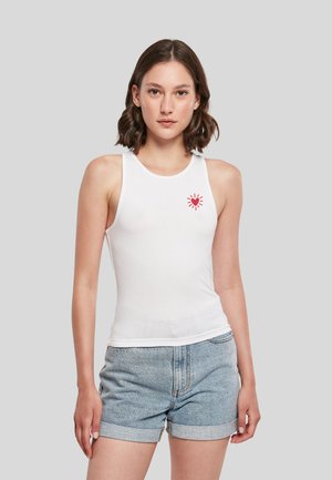 Jeune femme portant un haut blanc sans manches avec un symbole de cœur rouge sur la poitrine et un short en jean bleu clair, debout devant un fond gris.