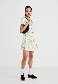 Witte bloemenjurk met kleurrijke planten; korte mouwen, uitlopende vorm. Geselecteerd met een zwarte leren tas, zwarte schoenen en witte sokken.