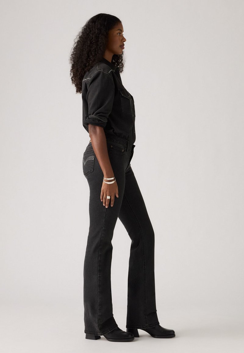 Une chemise en denim noir et un jean évasé présentent des coutures contrastantes. Le mannequin se tient de côté, portant des bottes à talons noires et des accessoires en argent.