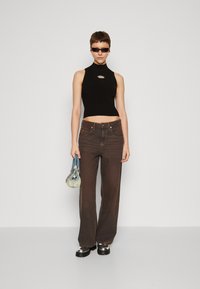 Schwarzes, geripptes, ärmelloses Top mit einem Ausschnitt, kombiniert mit braunen Weitenjeans. Das Model hält eine kleine blaue Handtasche und trägt eine Sonnenbrille.