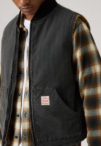 Črn denim telovnik z pokončnim ovratnikom, stranskimi žepi in rdečo tekstilno etiketo, položen preko kariraste flanelaste srajce v rjavih in zelenih odtenkih.