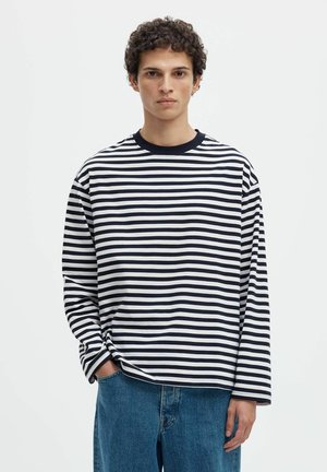 Jeune homme aux cheveux bouclés portant un t-shirt à manches longues rayé noir et blanc et un jean bleu, face à l’avant avec une main dans la poche.