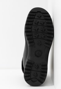Semelle de chaussure noire avec des crampons profonds, des motifs en forme d'étoile et rectangulaires, ainsi qu'un logo circulaire près du talon sur un fond blanc.