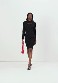 Robe noire à manches longues avec un décolleté ajouré, design ajusté et texture lisse. Associée à un petit sac à main rouge et des escarpins noirs.