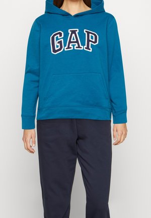 Person trägt blauen GAP-Hoodie mit Kängurutasche und dunkelblaue Hose, steht vor einfachem hellem Hintergrund.