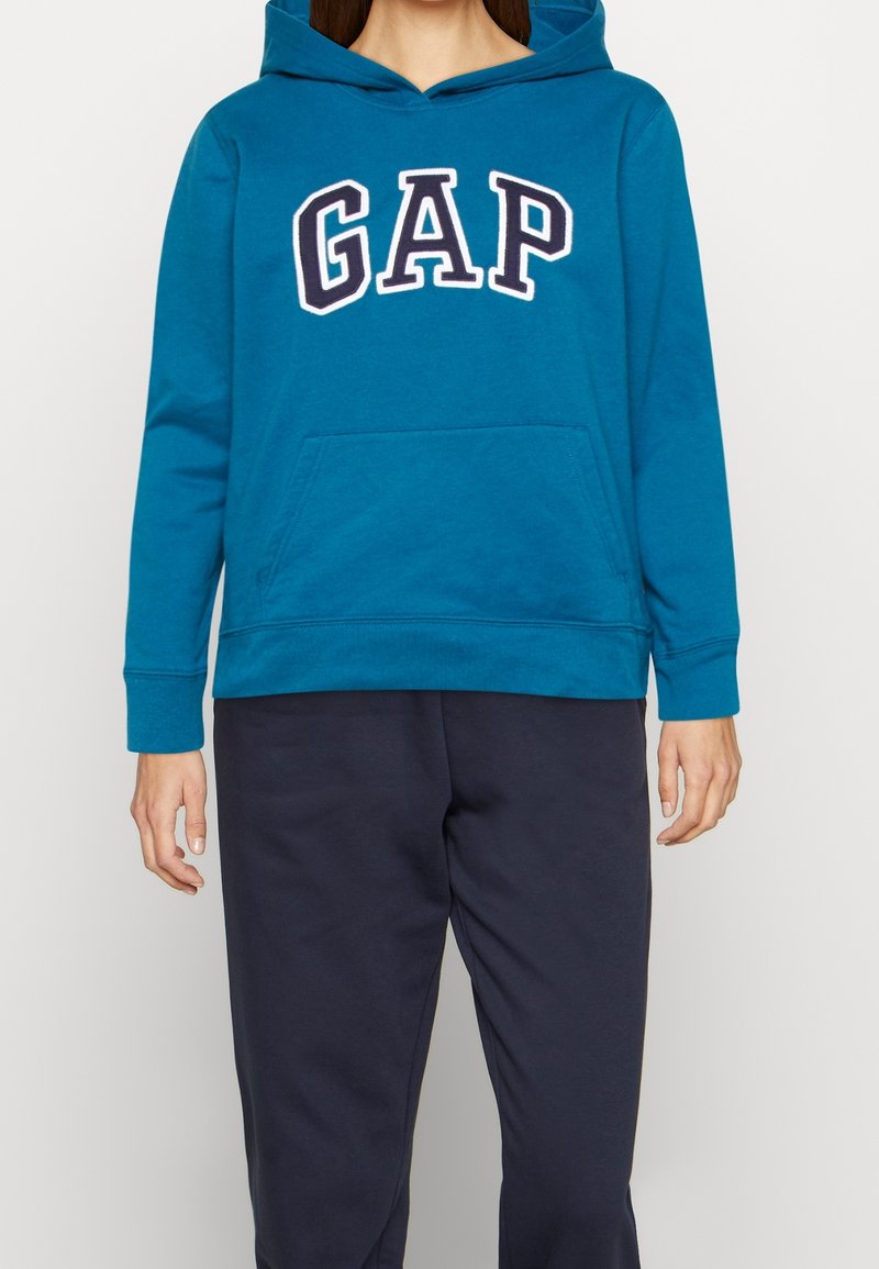 Personne portant un sweat à capuche bleu GAP avec poche kangourou et pantalon bleu marine, debout devant un fond clair uni.