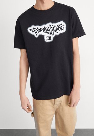 Czarna bawełniana koszulka z białym logo w stylu sprayu i prostokątną metką na środku. Krótkie rękawy, luźny krój.