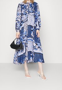 Lange blauwe jurk met patroon en ballonmouwen, met bloemen- en ruitontwerpen. Gecombineerd met een zwarte handtas en doorzichtige sandalen met hak.