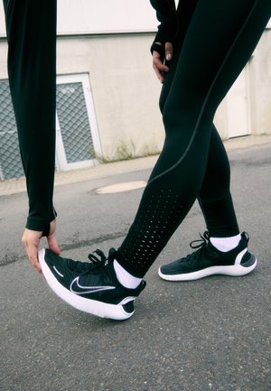 Sapatilhas desportivas Nike em preto com solas brancas, parte superior texturizada e acentos refletivos, combinados com leggings de compressão pretas com perfurações.