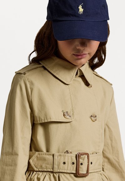 Beige dobbeltradet trenchcoat med belte i midjen, stor krage og glansfulle knapper. Marineblå cap med logo på forsiden.