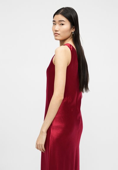 Νεαρή γυναίκα με μακριά σκουρόχρωμα μαλλιά, ντυμένη με αμάνικο, κόκκινο σατινέ φόρεμα, στέκεται και κοιτάζει πάνω από τον ώμο της πάνω σε απλό φόντο.