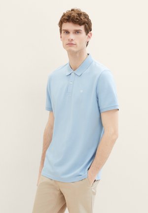 Lichtblauwe polo gemaakt van zacht materiaal met een platte gebreide kraag, twee knopen en een klein logo op de borst. Gecombineerd met beige broek.