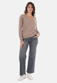 Pullover beige a scollo a V con polsini a coste, abbinato a jeans blu a vita alta e sneakers bianche. L'outfit presenta una vestibilità casual e una consistenza morbida.