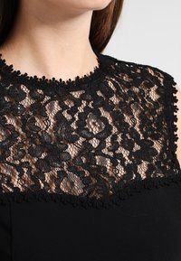 Zwarte jurk met een kanten halslijn met bloemmotieven en een geschulpte rand. Zachte stof met een aansluitende pasvorm en mouwloos ontwerp.