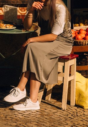 Ljusgrå satinklänning i midi-längd över en vit t-shirt, kombinerad med vita mesh-sneakers. Sittande på en träpall bredvid ett bord med bakverk.