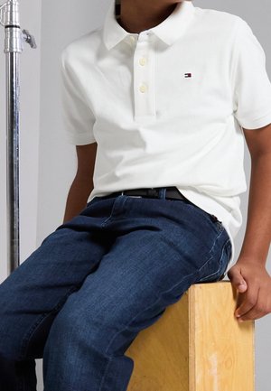 Kind trägt weißes Polohemd mit kleinem Logo, dunkelblaue Jeans und schwarzen Gürtel, sitzt auf einem Holzkasten vor einem schlichten Hintergrund.