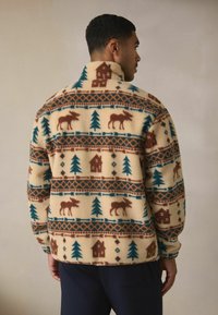 Kurtka polarowa z kremowym tłem, wzorem brązowych łosi, zielonych drzew i domów; wysoki kołnierz; miękka, teksturowana powierzchnia.