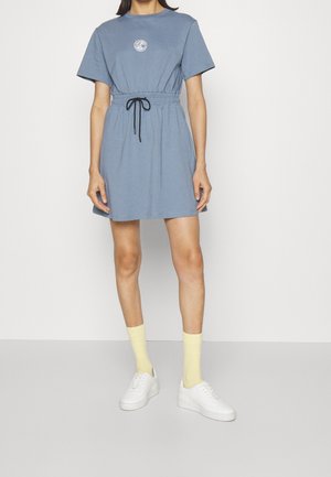 Kobieta ubrana w niebieską sukienkę z krótkim rękawem i czarnym paskiem ze sznurkiem w talii, żółte skarpetki i białe trampki, stojąca na jednolitym tle.