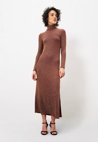 Langärmliges, Turtleneck-Kleid aus bronze-glänzendem Stoff, mit seitlichem Schlitz, figurbetonender Silhouette und fällt bis zu den Knöcheln. Getragen mit schwarzen Absatzschuhen.