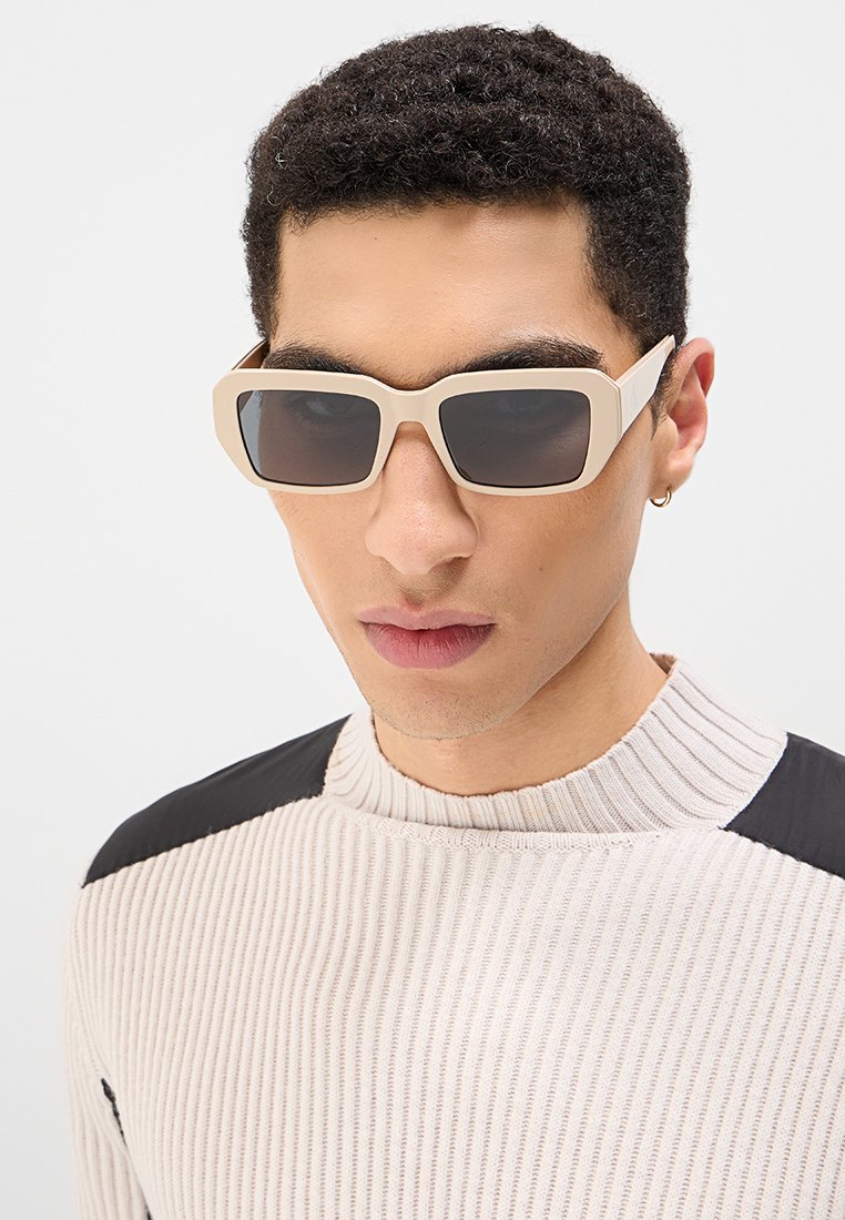 Beżowe okulary przeciwsłoneczne z geometrycznym wzorem i ciemnymi soczewkami. Noszone przez osobę w teksturowanym kremowym swetrze z czarnymi akcentami.