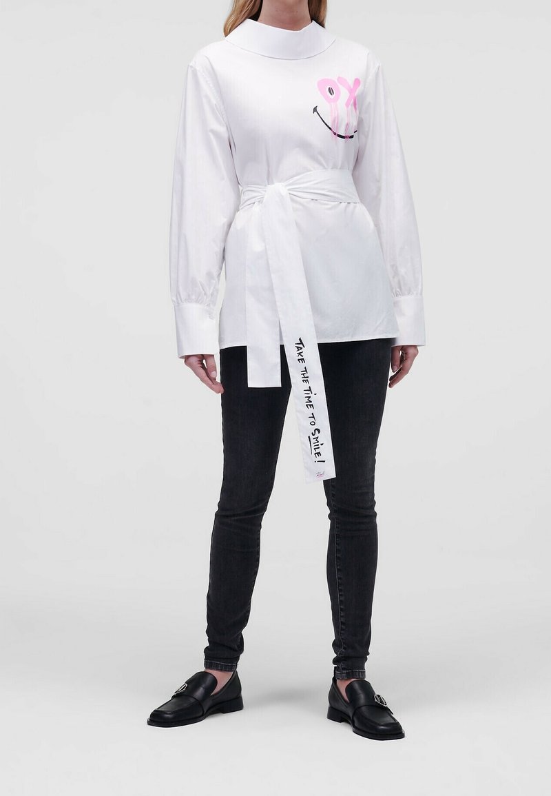 Osoba ubrana w białą bluzkę z różowym uśmiechniętym buźką i paskiem z napisem „Take the time to smile!” w połączeniu z czarnymi obcisłymi dżinsami i czarnymi mokasynami.