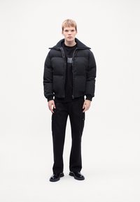 Black puffer jacket with high collar, zip closure, and horizontal quilted design; worn over a black sweater and paired with black cargo pants.