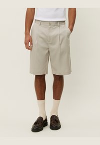 Beige plisserte shorts med knappelukking, sidelommer og en avslappet passform, kombinert med kremfargede ribbestrikkede sokker og mørkebrune sko.