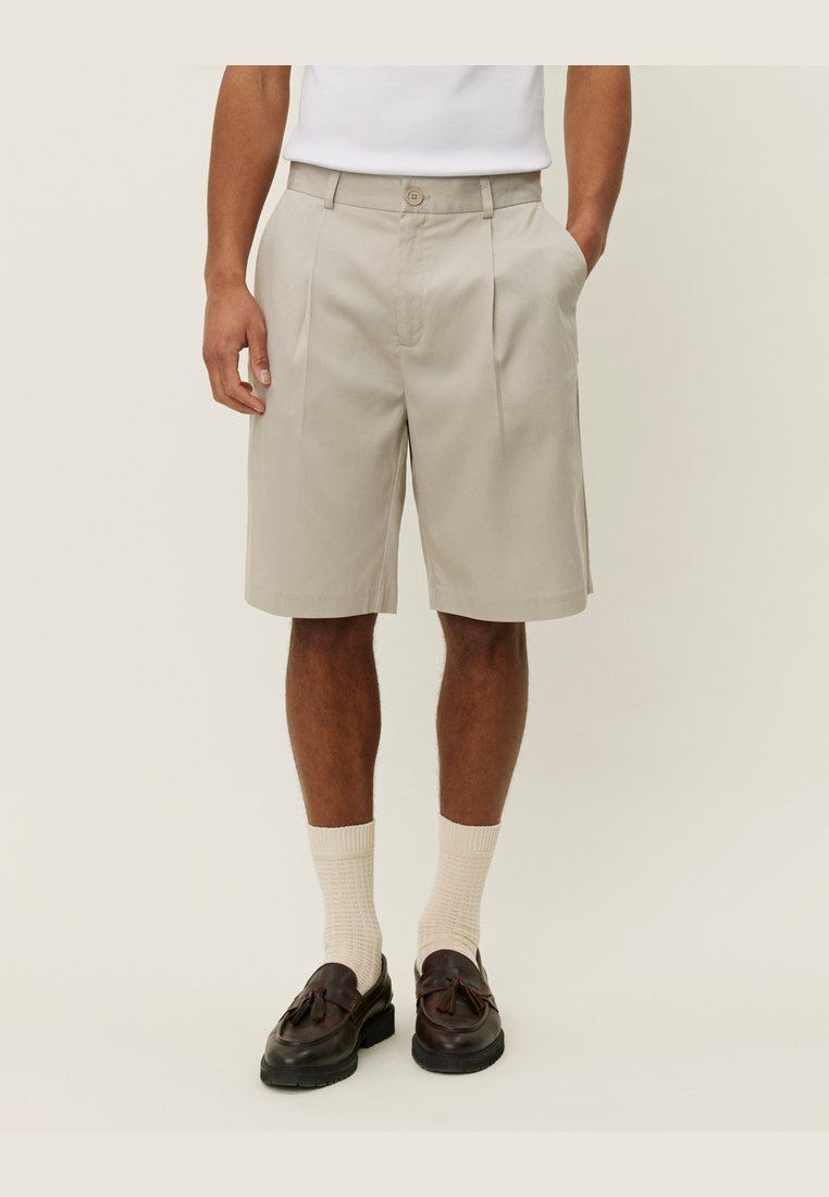 Beige plisserede shorts med knaplukning, sidelommer og en afslappet pasform, parret med cremefarvede ribbede sokker og mørkebrune sko.