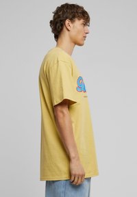 Gul bomulds t-shirt, afslappet pasform med korte ærmer, prydet med et farverigt grafisk design på forsiden. Blød tekstur med ensfarvet look.
