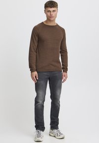Pulover lung maro cu mânecă lungă, guler rotund și manșete cu nervuri, asociat cu blugi slim-fit gri și adidași gri deschis cu accente texturate.