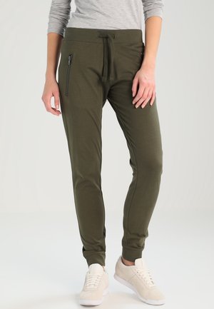Dopasowane zielone spodnie jogger w kolorze oliwkowym z kieszeniami na zamek po bokach, noszone z beżowymi trampkami i jasnoszarym koszulą z długim rękawem.