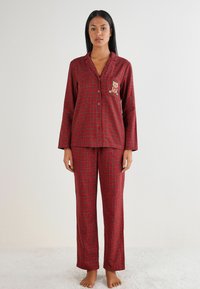 Rød ternet pyjamas sæt, der består af en skjorte med knaplukning og lange ærmer samt en bjørn broderet på brystlommen, kombineret med løstsiddende bukser.
