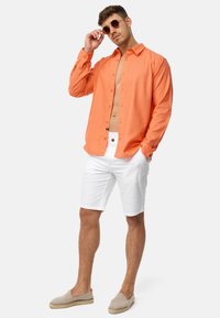 Orange langærmet skjorte, uoffentliggjort øverst, parret med hvide shorts og beige slip-on sko. Modellen bærer runde solbriller.