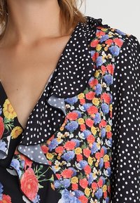 Blouse à motifs floraux et à pois, présentant un fond noir, un col en volants et des couleurs vives, y compris des fleurs rouges, bleues et jaunes.