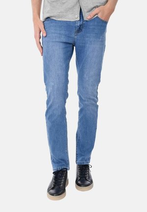 Uomo che indossa jeans blu aderenti, camicia grigia e sneakers nere con lacci, in piedi con una mano in tasca su uno sfondo bianco.