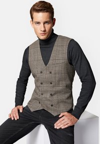 Brązowa kamizelka w kratę z podwójnym zapięciem, kontrastowy czarny golf i czarne dżinsy. Gładka faktura i dopasowany krój.