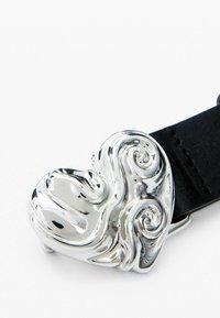 Fibbia a forma di cuore in argento con motivi intrecciati, fissata a una cinghia in pelle nera. Texture metallica liscia e finitura lucida.