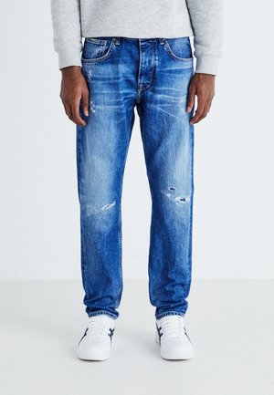 Jeans de mezclilla azul oscuro con corte recto, que presentan un ligero desgaste y un lavado deslavado. Combinados con zapatillas blancas.