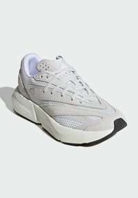Zapatilla atlética de color gris claro con parte superior de malla, acentos texturizados y cordones tonales. Suela intermedia blanca gruesa con suela exterior negra; presenta la marca en el costado.