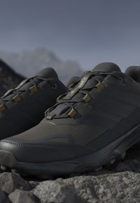 Chaussures de trail running gris foncé avec semelles robustes reposant sur un terrain rocheux sous un ciel nuageux.