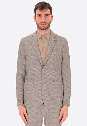 Uomo che indossa un completo beige con trama e bottoni coordinati su camicia marrone chiaro con colletto, in piedi davanti a uno sfondo bianco semplice.