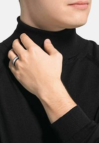 Czarny sweter z golfem o prążkowanej fakturze, noszony przez osobę delikatnie trzymającą się za szyję. Na ich dłoni widoczny jest srebrny i czarny pierścionek.