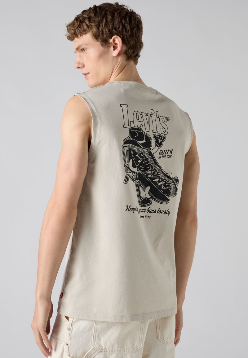 Homme portant un débardeur beige Levi's avec un motif noir représentant un hot-dog allongé sur une chaise et le texte « Keep your buns toasty » au dos.