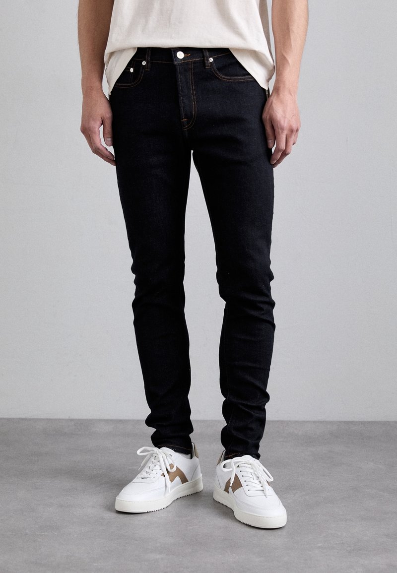 Mand iført slanke sorte jeans og hvide sneakers, der står på en grå gulv mod en ensfarvet grå væg.